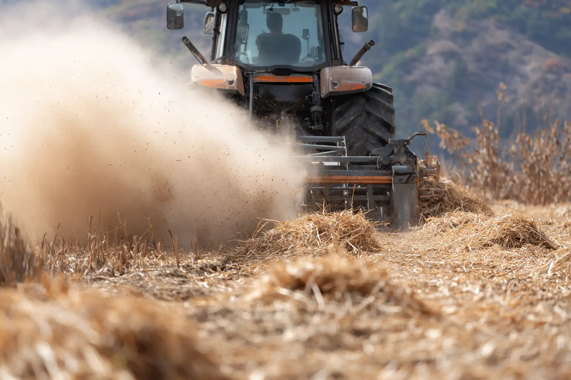 side view of a tractor using a vertical drum mower on dry grassland, visible dust cloud rising behind the machine
